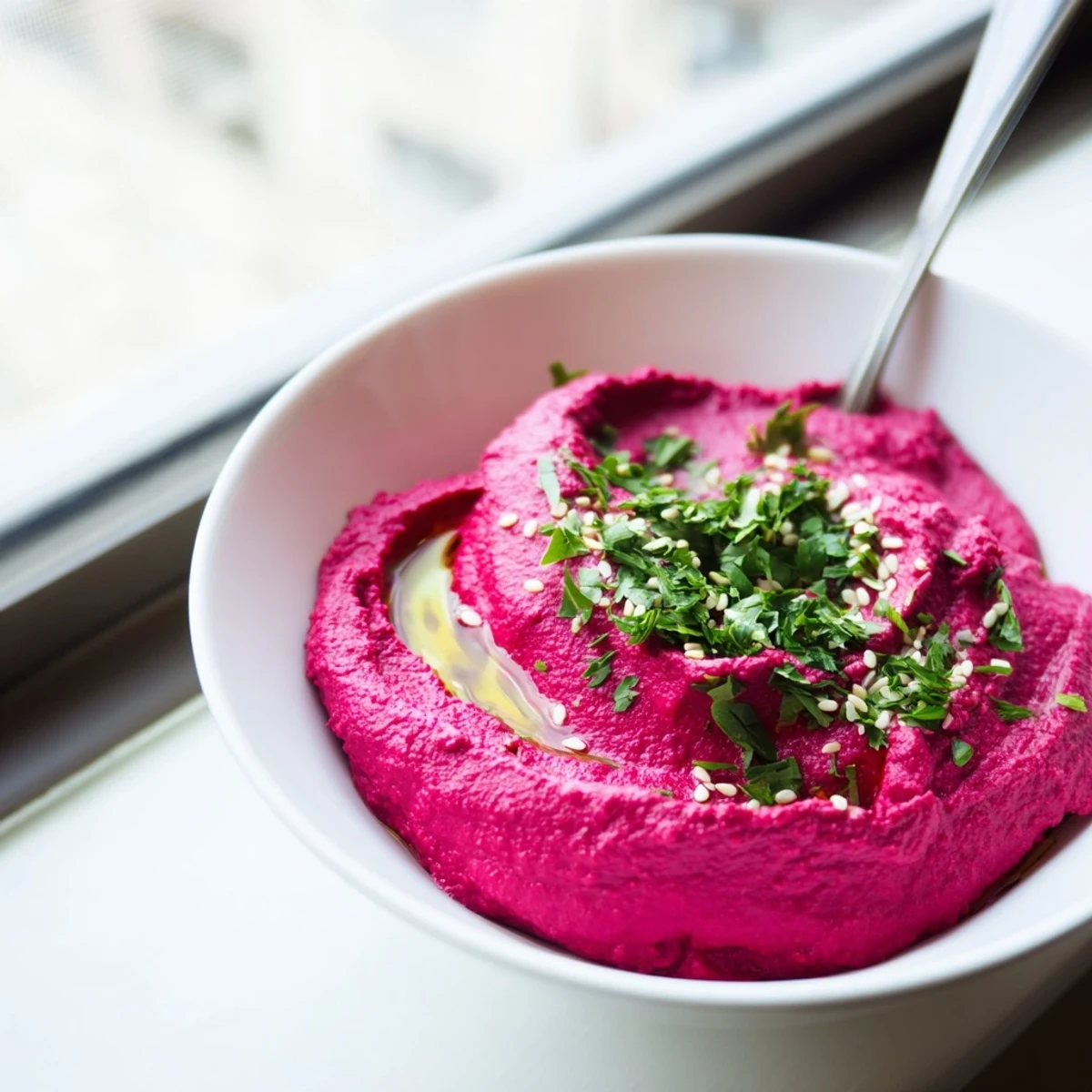 Creamy roasted beet hummus in a white bowl, garnished with olive oil drizzle and parsley, served with pita chips.  
