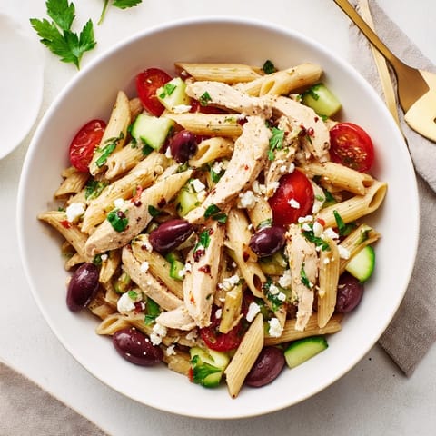 Tender slices of lemony grilled chicken rest on a vibrant pasta bowl with crisp cucumbers, red onions, and briny olives.